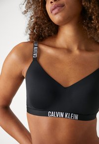 Crni bralette od gladkog materijala, s V-izrezom i podešavnim trakama na kojima je bijelim slovima ispisano "CALVIN KLEIN".