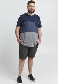 Kurzärmeliges, zweifarbiger T-Shirt in Marineblau, Blau und Grau mit einer Brusttasche, kombiniert mit olivgrünen Shorts und schwarzen Turnschuhen.
