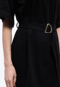 Μαύρο φόρεμα με υφασμάτινη ζώνη που έχει λαμπερό χρυσό τριγωνικό αγκράφα, φορεμένο από ένα άτομο με ορατό αριστερό μπράτσο σε λευκό φόντο.