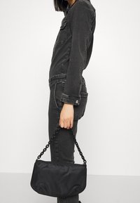 Pochette noire texturée avec poignée en chaîne, dotée d'une fermeture éclair et d'une forme arrondie et douce. Portée avec une tenue sombre assortie.