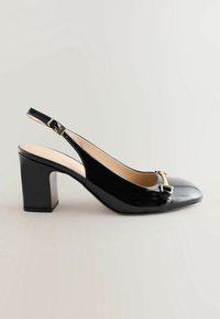 Chaussure slingback en cuir verni noir avec talon bloc et boucle dorée sur l’orteil, présentée sur un fond uni.