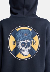 Felpa con cappuccio blu navy con un grande motivo grafico di un teschio con un berretto blu, un occhio rosso e uno sfondo giallo, incorniciato da un design circolare.