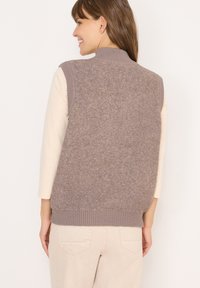 Brauner Strickwesten mit einem hohen Kragen, strukturiertem Rücken und geripptem Saum. Trägt eine helle Langarmbluse und kombiniert mit beigen Hosen.