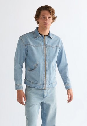 Jeune homme aux cheveux bouclés portant une veste en jean bleu clair zippée et un jean assorti, debout devant un fond blanc uni.