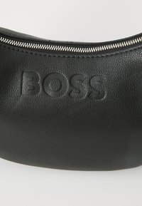 Bolso de cuero negro con un diseño curvado, superficie texturizada, logo en relieve y un cierre de cremallera en tono plateado en la parte superior.