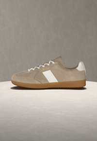 Bež superga iz semiša z belimi stranskimi črtami, belimi vezalkami, belim jezičkom na peti in gumijastim podplatom, označena z napisom "Polo" na strani blizu vezalk.