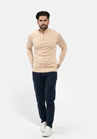 Beige Zip-Kragen Langarmoberteil kombiniert mit navy Jogginghosen. Nennenswerte Details sind gerippte Bündchen und ein lockerer Schnitt. Weiße Sneaker runden den Look ab.