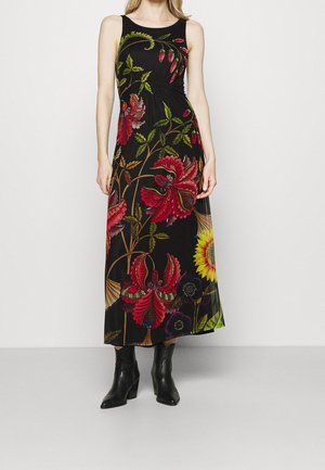 Kobieta ubrana w czarną, bezrękawnikową suknię maxi z dużymi czerwonymi, żółtymi i zielonymi motywami kwiatowymi, w połączeniu z czarnymi botkami na obcasie.