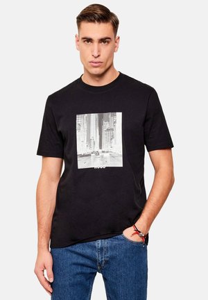 Młody mężczyzna z krótkimi włosami nosi czarną koszulkę z czarno-białym pejzażem miejskim oraz niebieskie dżinsy, z ręką w kieszeni i neutralnym wyrazem twarzy.