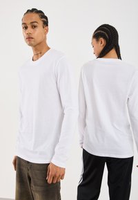 Deux personnes avec des cheveux tressés, faisant face dans des directions opposées, portant des chemises blanches à manches longues ; l'une visible de face, l'autre de dos.