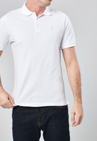 Homme portant un polo blanc à manches courtes avec un petit logo brodé sur la poitrine et un jean bleu foncé.
