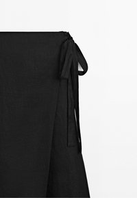 Gros plan d'une jupe noire en tissu avec un détail de nœud sur le côté, présentée sur un fond blanc uni. Aucune personne ou autre objet visible.