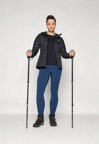 Jauna sieviete, tērpusies melnā pūkainajā jakā, zilos legingos un pārgājienu zābakos, stāv, turot divus melnus pārgājienu nūjas pret platu fona.