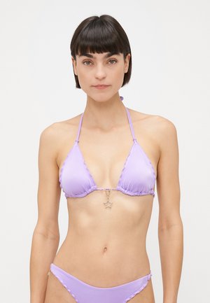 Woman with short dark hair wearing a lilac bikini with a gold star charm on the top, standing against a plain light background.