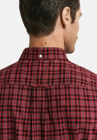 Red and black plaid shirt featuring a button-down collar, pleats at the back, and a smooth texture. White button detail at the collar.