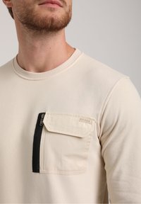 Sudadera beige con cuello redondo, que cuenta con un bolsillo en el pecho izquierdo con un cierre negro y una solapa. Tejido de algodón suave con detalles acanalados.