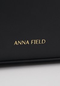 Sort syntetisk lædertaske med prægede "ANNA FIELD" logo i guld. Glat overflade med rene linjer og ingen synlige mønstre.