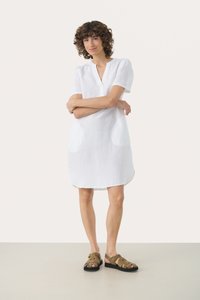 Weißes Leinenkleid mit kurzen Ärmeln, einem V-Ausschnitt und zwei runden Taschen. Der Saum ist leicht geschwungen, kombiniert mit braunen Riemchensandalen.