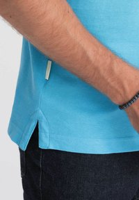 Vaaleansininen polo-paita, jossa on tekstuuri, sivureikä ja pieni etiketti helmassa, yhdistettynä tummiin farkkuihin.