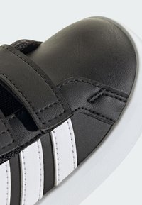 Černý kožený teniska s dvěma bílými proužky, kulatou špičkou a páskem na suchý zip přes střední část chodidla pro bezpečné zapnutí.