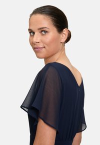 Robe bleu marine avec des manches flottantes transparentes, dos ouvert et fermeture éclair discrète. Texture lisse, taille ajustée et silhouette élégante.