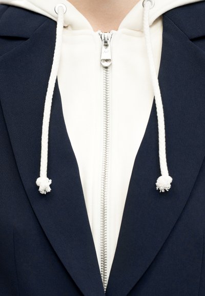 Close-up of a white zip-up hoodie with drawstrings worn under a dark navy blazer.