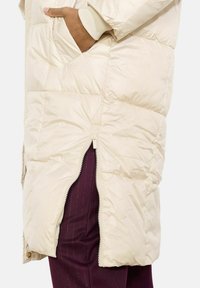 Długi beżowy kamizelka puchowa z pikowanym wzorem, bocznymi kieszeniami i zamkiem błyskawicznym z przodu. Nałożona na ciemnofioletowe spodnie z teksturą prążków.