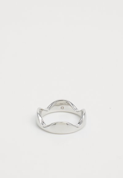 Anello in argento lucido e levigato con un design ondulato e sinuoso posato su una superficie bianca liscia.