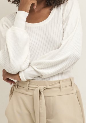 Vrouw draagt een witte geribde trui met lange mouwen en opgeblazen mouwen, gecombineerd met beige broek met hoge taille en een stoffen riem.