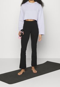 Sweatshirt court lavande à manches larges associé à des leggings évasés noirs. Tenant une bouteille d'eau rose. Debout sur un tapis de yoga gris.