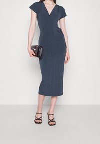 Houtskoolgrijze, middellange wikkeljurk met korte mouwen en een strikdetail in de taille. Model houdt een zwarte handtas vast en draagt zwarte sandalen met bandjes.