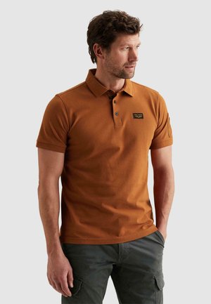 Muž s krátkymi kučeravými vlasmi, oblečený v hnedej krátkosrktej polo košeli a tmavých cargo nohaviciach, pozerá sa doprava s rukami uvoľnenými pozdĺž tela.