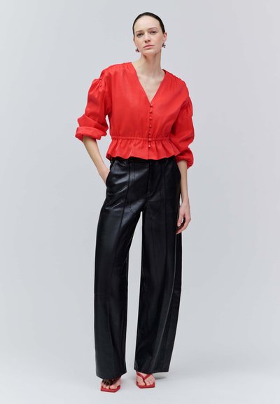 Kobieta stoi, ubrana w czerwoną, zapinaną na guziki bluzkę z podwiniętymi rękawami, czarne szerokie spodnie ze skóry oraz czerwone buty na obcasie z odkrytymi palcami na jasnoszarym tle.