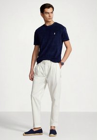 Μπλε σκούρο κοντομάνικο t-shirt, λευκό παντελόνι σε άνετη γραμμή και μπλε εσπαντρίγιες. Το t-shirt έχει μικρό λογότυπο στο στήθος.