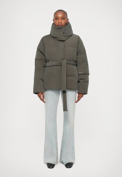 Une personne se tient face à l'avant, portant une veste grise rembourrée ceinturée, un jean bleu clair évasé et des chaussures noires sur un fond blanc.