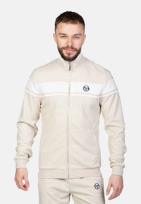 Giacca da track beige con strisce bianche, colletto alto, zip completa e logo. Texture morbida, design aderente, pantaloni abbinati.
