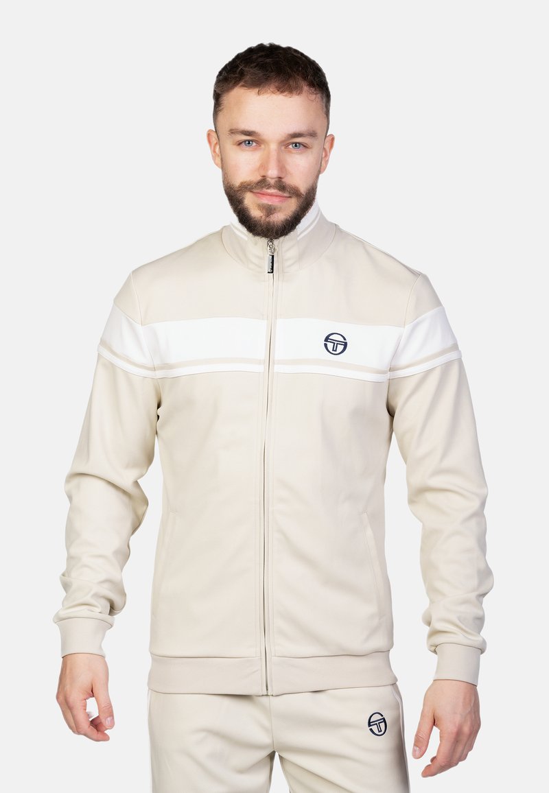 Giacca da track beige con strisce bianche, colletto alto, zip completa e logo. Texture morbida, design aderente, pantaloni abbinati.
