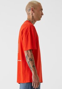 Oransje bomull t-skjorte med korte ermer, løs passform. Har tekstdesign på brystet og ryggen. Brukes med blå denimjeans.