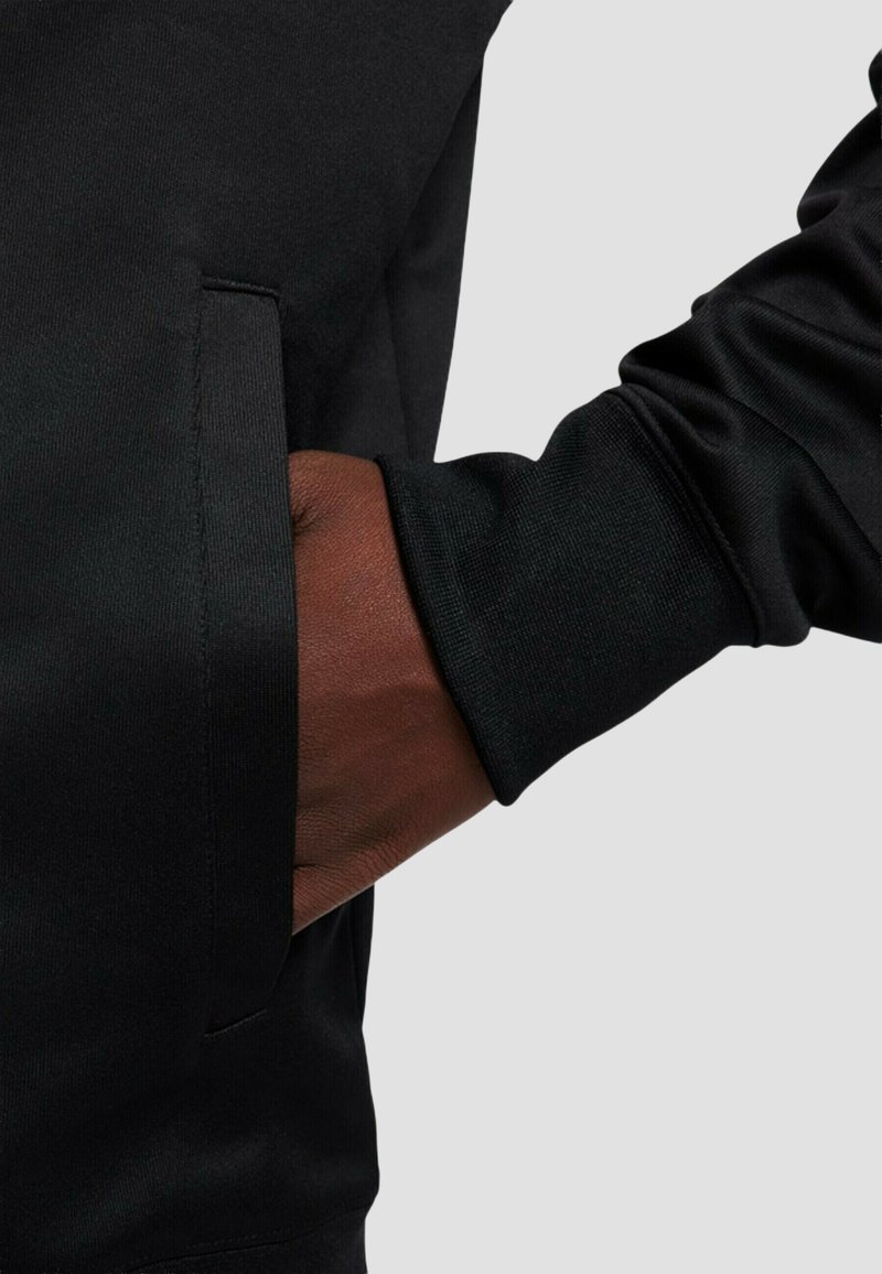 Mano con tono de piel oscuro colocada dentro del bolsillo de una prenda negra de manga larga sobre un fondo claro y liso.