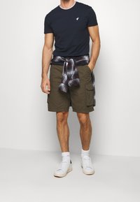 Marinblå crewneck t-shirt med vit krage, olivgröna cargoshorts med sidofickor och vita sneakers. Rutig flanellskjorta knuten i midjan.