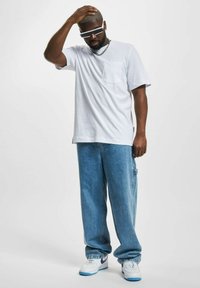 T-shirt bianco a maniche corte con taschino sul davanti, abbinato a jeans baggy di denim azzurro chiaro. Gli accessori includono occhiali da sole e una collana in argento.