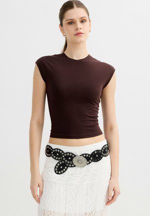 Joven mujer con una camiseta sin mangas ajustada de color marrón oscuro y una falda blanca de encaje con un cinturón ancho negro que tiene ojales metálicos y una hebilla grande.