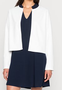 Giacca bianca cropped con collo rotondo, caratterizzata da maniche lunghe e senza chiusure, indossata su un abito blu navy con scollatura a V.