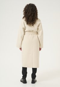 Palton crem cu garnitură, cu mâneci lungi, de lungime medie până la gambe, având o croială dreaptă și o textură moale. Combinație cu pantaloni negri cu croială dreaptă și pantofi.