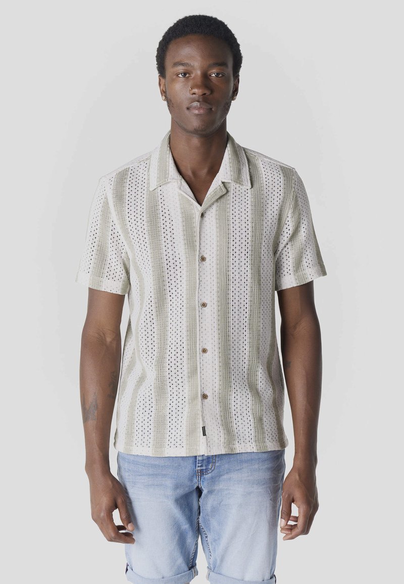 Jeune homme portant une chemise en maille à manches courtes, boutonnée, rayée beige et blanche, et un short en denim bleu clair, debout devant un fond uni.