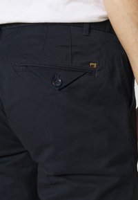 I pantaloni di cotone blu navy presentano una tasca laterale con chiusura a bottone e un piccolo logo dorato. Il tessuto ha una texture liscia e semplice.