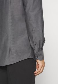 Chemise gris foncé avec un motif de petits points subtils, manches longues avec poignets à boutons, et un ourlet courbé. Le tissu semble lisse et respirant.