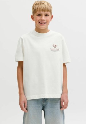 Lyshåret dreng smilende, iført en løs hvid T-shirt med lille trykt logo på brystet og lyseblå denimjeans, stående mod en ensfarvet baggrund.