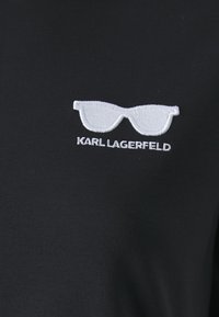 Juodas marškinėliai su balta siuvinėta akinių grafiką ir tekstu "KARL LAGERFELD" apačioje. Minkšta, lygi tekstūra ir klasikinis kirpimas.
