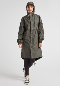 Impermeable de color verde oliva con capucha, cremallera completa, puños elásticos y un parche con logo en la manga; combinado con leggings negros y zapatillas.